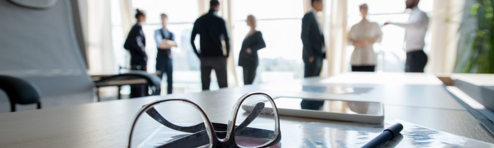 Close-up of conference table with personal stuff such as glasses, financial paper, pen and smartphone, business people talking in background
